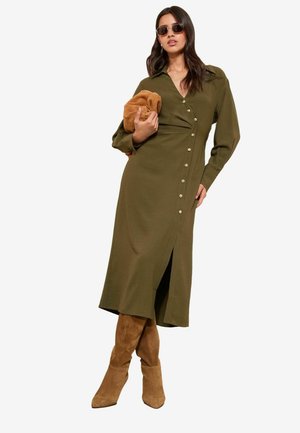 Olivgrünes langärmeliges Kleid mit durchgehender Knopfleiste und seitlichem Schlitz. Kombiniert mit braunen kniehohen Wildlederstiefeln und einer braunen Clutch in der Hand.