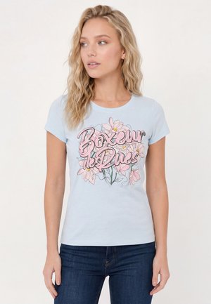 Femme aux longs cheveux blonds ondulés portant un t-shirt bleu clair avec un motif floral rose et un texte, associé à un jean bleu foncé.