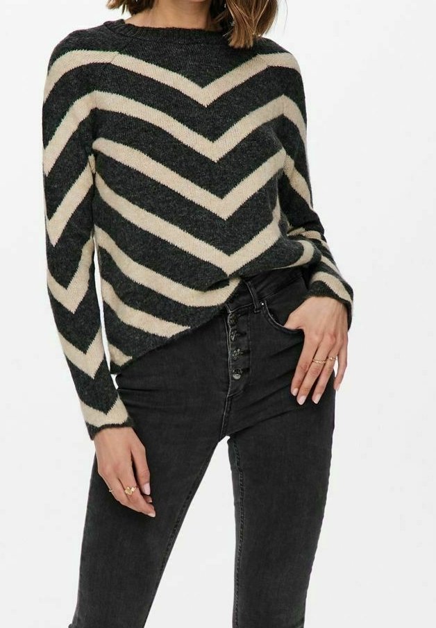 Femme portant un pull à rayures en chevron noir et beige, rentré dans un jean noir taille haute à boutons, main posée dans la poche.