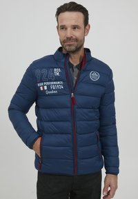 Mėlynas pūkinis švarkas su siuvinėjimu, raudonais užtrauktuko akcentais ir spausdintomis grafika. Aukšta apykaklė, šoninės kišenės ir prigludusi forma.