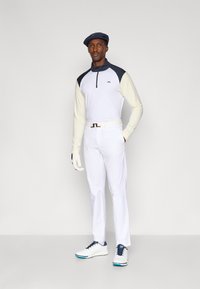 Polo branco de mangas compridas com detalhes em azul-marinho, emparelhado com calças brancas e um cinto. Usado com um boné azul-marinho e sapatos de golfe brancos, com detalhes azuis.