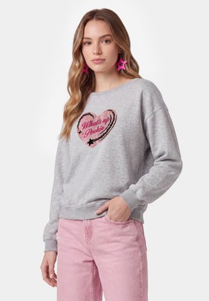 Jonge vrouw met lang golvend haar draagt een grijze trui met een roze hart en de tekst "What's up Pookie" en roze sterrenvormige oorbellen.