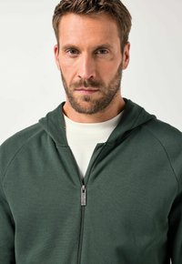 Grüner Zip-Hoodie aus strukturiertem Stoff, mit einer Kapuze mit Zugband und einem silbernen Reißverschluss. Über einem hellen Shirt getragen.