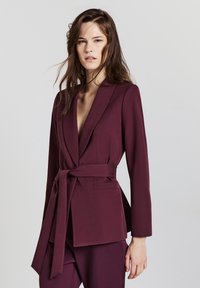 Blazer bordeaux con maniche lunghe, revers e una cintura in vita. Presenta due tasche frontali e un tessuto liscio e testurizzato.
