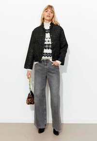 Veste noire texturée portée sur un pull blanc à motif géométrique, associée à un jean gris taille haute à jambes larges et à un petit sac à main imprimé léopard.
