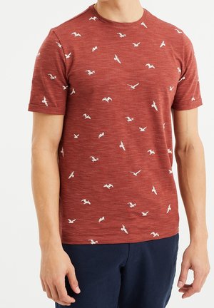 Mężczyzna w czerwonej koszulce z krótkim rękawem z białymi wzorami ptaków, w ciemnoniebieskich spodniach, stojący na białym tle.