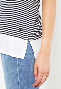 Camiseta a rayas con rayas azules y blancas, con un bajo blanco transparente y un pequeño logo bordado en la parte inferior izquierda. Combinada con jeans azules.