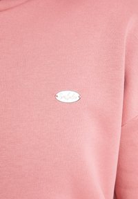 Sweatshirt en tricot rose avec une étiquette ovale argentée portant l'inscription "Sunlato", présentant une texture lisse et une coupe décontractée.