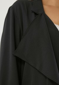 Collo di cappotto nero texturizzato su una persona che indossa un indumento bianco sottostante, mostrando i dettagli del tessuto e i diversi strati dell'abbigliamento.