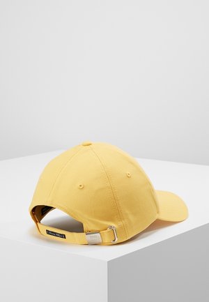 Casquette de baseball en coton jaune avec une sangle à boucle métallique réglable, posée sur une surface blanche d'angle contre un fond gris uni.