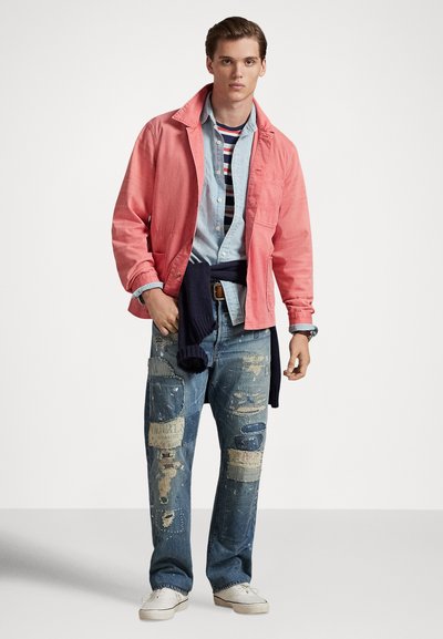 Jauns vīrietis valkā koraļļu krāsas jaku, svītrainu kreklu zem džinsa krekla, plāksteriem izlabotas džinsas, baltas sporta kurpes un tumši zilu džemperi, kas ir sasiets ap vidukli.