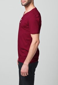 Burgundské tričko s krátkým rukávem a henley výstřihem, vybavené třemi knoflíky, vyrobené z měkké látky s hladkým povrchem.