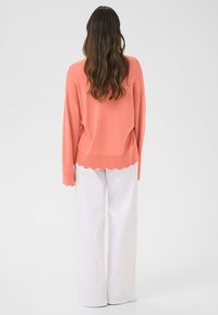 Koralni pleten pulover z valovitim robom, v kombinaciji z ohlapnimi belimi hlačami. Pulover ima spuščena ramena in sproščen kroj.