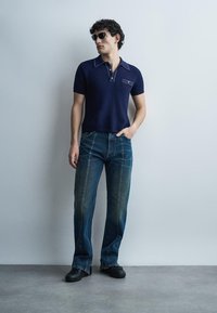 Άνδρας που φοράει σκούρο μπλε πλεκτό πόλο, μπλε τζιν με κατακόρυφες ραφές, μαύρα παπούτσια και γυαλιά ηλίου, στέκεται μπροστά σε ένα απλό ανοιχτό γκρι τοίχο.