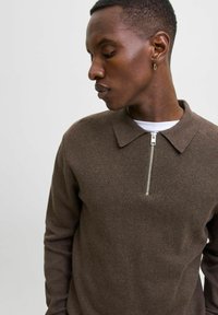 Muž v hnědém dlouhém pleteném polo s stříbrným předním zipem, dívající se dolů a vlevo na jednobarevném světlém pozadí.