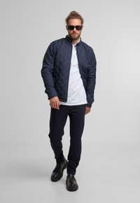 Marineblauwe gewatteerde bomberjack met een hoge kraag, gecombineerd met een witte polo en donkere broek, afgemaakt met zwarte boots met een grove zool.