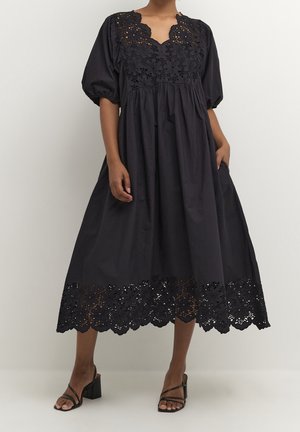 Robe en coton noir avec des manches bouffantes, ourlet en dentelle festonnée et motif floral découpé au niveau du col. Silhouette en A.