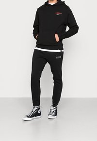 Felpa nera con tasca frontale e testo rosso, abbinata a pantaloni jogger neri con vita bianca e branding, indossata con sneaker nere alta.