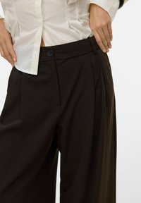 Brune bukser med brede ben og en glat tekstur, med forreste læg og en enkelt sort knaplukning ved taljen.