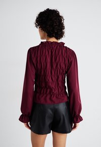 Μπορντό μπλούζα με σχεδίαση καπνιστής πλάτης, μακριά μανίκια και συγκεντρωμένα μανίκια, συνδυασμένη με μαύρες δερμάτινες σορτς. Λείος υφασμάτινος ιστός.