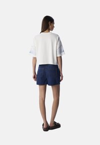Witte cropped t-shirt met blauwe grafische details op de mouwen, gecombineerd met blauwe denim shorts. Model draagt zwarte plataformasandalen.