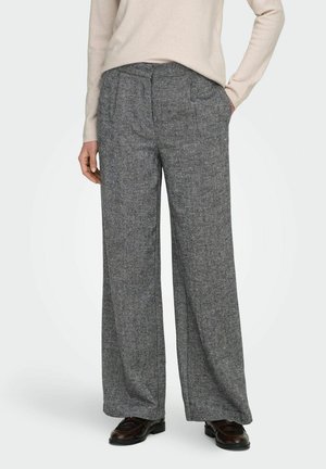 Femme portant un pantalon large grise taille haute associé à des mocassins marron foncé et un haut beige à manches longues, une main dans la poche.