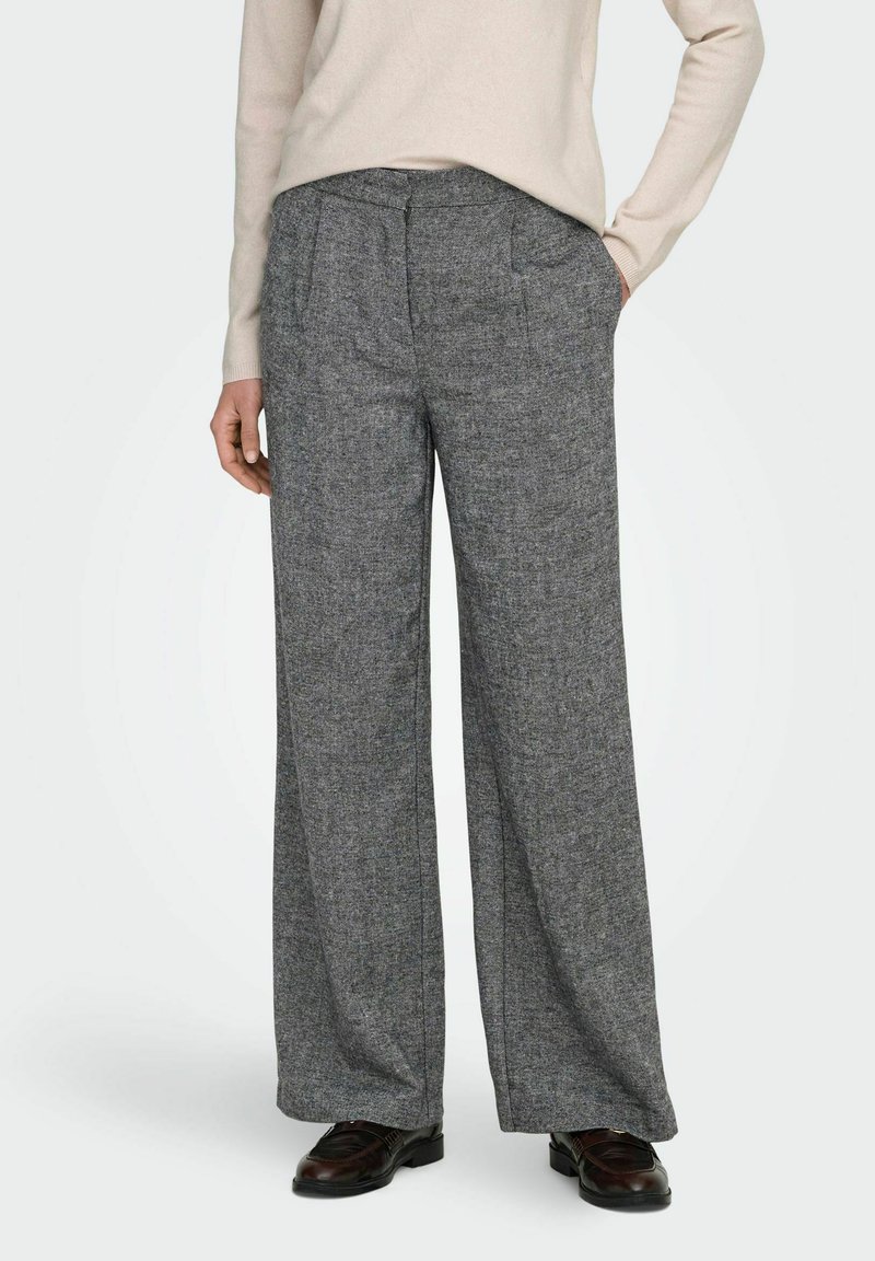 Moteris vilki pilką aukšto liemens, plačių kojų kelnių, derinamų su tamsiai rudos spalvos loafers tipo bateliais ir šviesiai smėlio spalvos ilgomis rankovėmis marškiniais, viena ranka į kišenę.