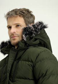 Groene pufferjas met een capuchon, voorzien van een afneembare nepleer trim, ritssluitingen en een doorgestikt ontwerp. De jas heeft een solide, gladde textuur.