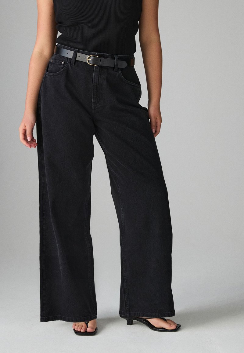 Personne portant un jean noir taille haute à jambes larges avec ceinture, un top noir sans manches et des sandales à talons ouvertes noires, debout sur un sol gris.