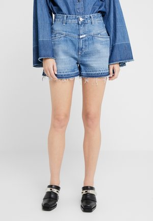 Personne portant un short en denim bleu moyen avec des ourlets effilochés, une chemise en denim bleu à manches longues avec des manches larges, et des chaussures slip-on noires avec des chaînes argentées.