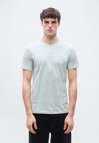 Νέος άνδρας με σγουρά μαλλιά, φορ ώντας ανοιχτό γκρι κοντομάνικη μπλούζα με στρογγυλή λαιμόκοψη και μαύρο παντελόνι, στέκεται μπροστά σε απλό λευκό φόντο.