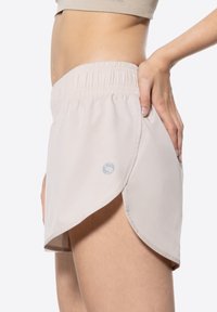 Pantaloni sportivi di un colore beige chiaro con vita elastica, design con spacco laterale e un logo discreto sulla coscia sinistra. Materiale liscio e traspirante.