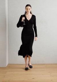 Robe en tricot côtelé noir à manches longues, décolleté en V profond et détails de boutons. Associée à des ballerines noires et un petit sac à main. Fond en bois.