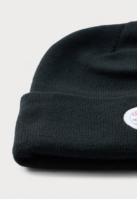 Zwarte gebreide beanie met opgevouwen rand, voorzien van een kleine ronde geborduurde patch met rode en blauwe tekst op een witte achtergrond.