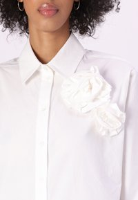 Camicia di cotone bianca con colletto classico e due decorazioni a forma di rosa in tessuto sul lato sinistro. Texture liscia e chiusura con bottone.