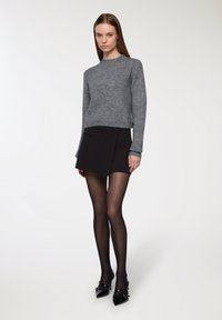 Kobieta stoi w szarej dzianinowej bluzie, czarnej mini spódnicy, przezroczystych czarnych rajstopach oraz czarnych lakierowanych szpilkach z paseczkami.