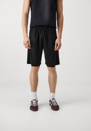 Ein Mann trägt schwarze knielange Shorts mit roten Seitenstreifen, ein schwarzes ärmelloses Shirt, weiße Socken und dunkle Sneakers und steht auf einem weißen Boden.