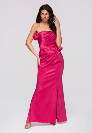 Kobieta w bezramkowej satynowej sukni w kolorze fuksji z drapowanymi detalami i boczną kokardą, pozująca z jedną ręką przy włosach, a drugą na biodrze.