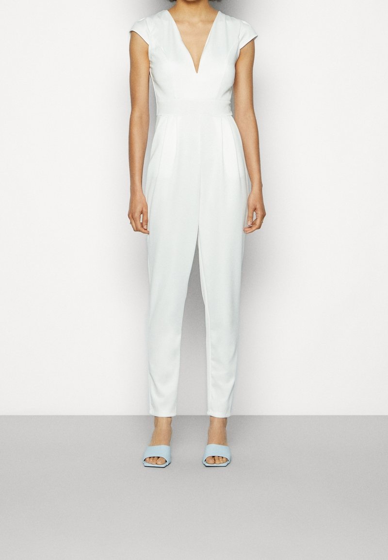 Mono blanco con mangas tipo cap, escote en V profundo y plisados. Combinado con mules de apertura delantera en azul claro. Textura suave, silueta ajustada.