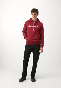 Rote Baumwollkapuzenjacke mit weißem "CHAMPION"-Logo, vordertasche, verstellbare Kordelzüge. Kombiniert mit schwarzen Cargo-Hosen und schwarzen Schuhen.