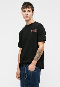 Zwart katoenen t-shirt met korte mouwen en een ronde hals, met een rood-wit logo op de linkerkant van de borst. Casual fit.