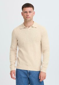Pullover beige lavorato a maglia con scollatura a V e colletto corto, dotato di maniche lunghe e polsini a coste. Abbinato a jeans in denim blu.