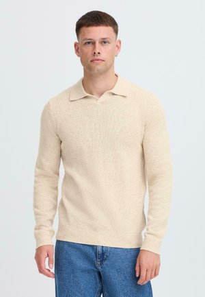 Suéter de punto beige con escote en forma de V y cuello corto, que presenta mangas largas y puños acanalados. Combinado con jeans de mezclilla azules.