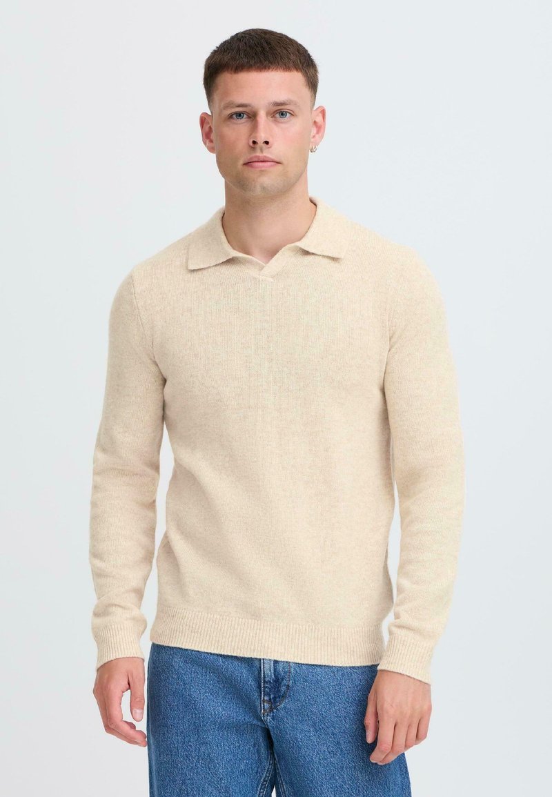 Pullover beige lavorato a maglia con scollatura a V e colletto corto, dotato di maniche lunghe e polsini a coste. Abbinato a jeans in denim blu.