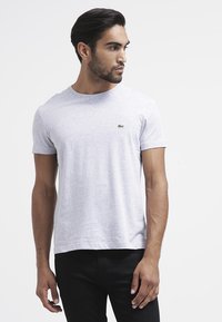 Grijze korte mouwen t-shirt van katoen, met een ronde halslijn en een klein groen logo op de linkerborst. Draagt met zwarte broek.