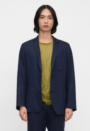 Jong persoon met halflang zwart haar, gekleed in een marineblauw colbert over een olijfgroen shirt, tegen een egale lichte achtergrond.