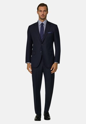 Mand iført en mørk navy jakkesæt med en blå mønstret skjorte, navy slips med hvide prikker, matchende lommetørklæde og sorte dress-sko.