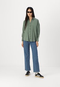 Grønn skjorte med knapper og avslappet passform, blå vide jeans og svarte sneakers med hvite striper; har lange ermer og en jevn tekstur.