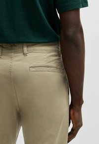 Chinos beige con una textura suave, que cuentan con un bolsillo trasero y una pequeña etiqueta de logo. El portador lleva una camisa verde oscura y un brazo visible.