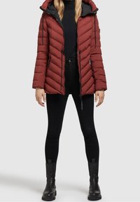 Chaqueta acolchada roja con capucha, que presenta un acolchado en forma de flecha, cremallera frontal y lazadas. Combinada con jeans ajustados negros y botines.